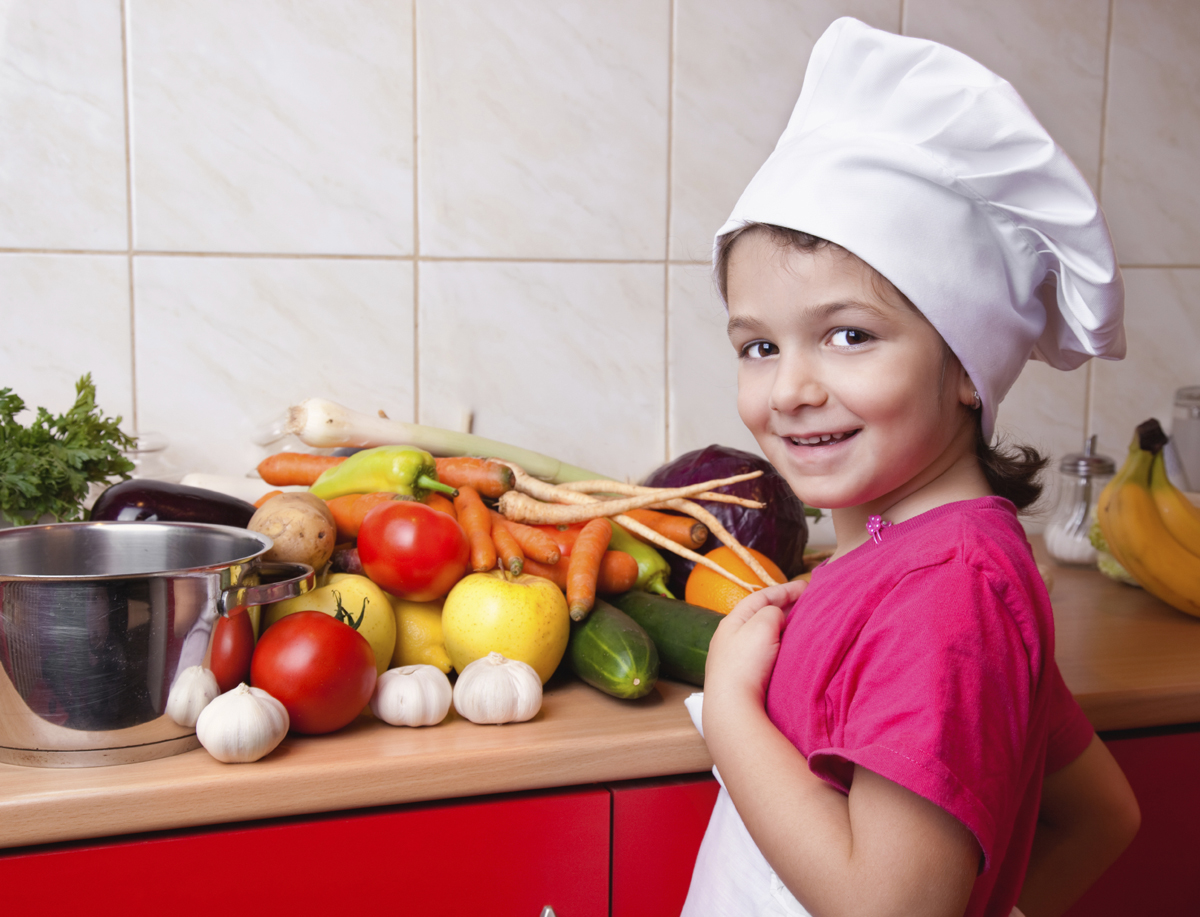 Youth wearing a chef hat stands beside a kitchen counter filled with fresh fruits and vegetables.
